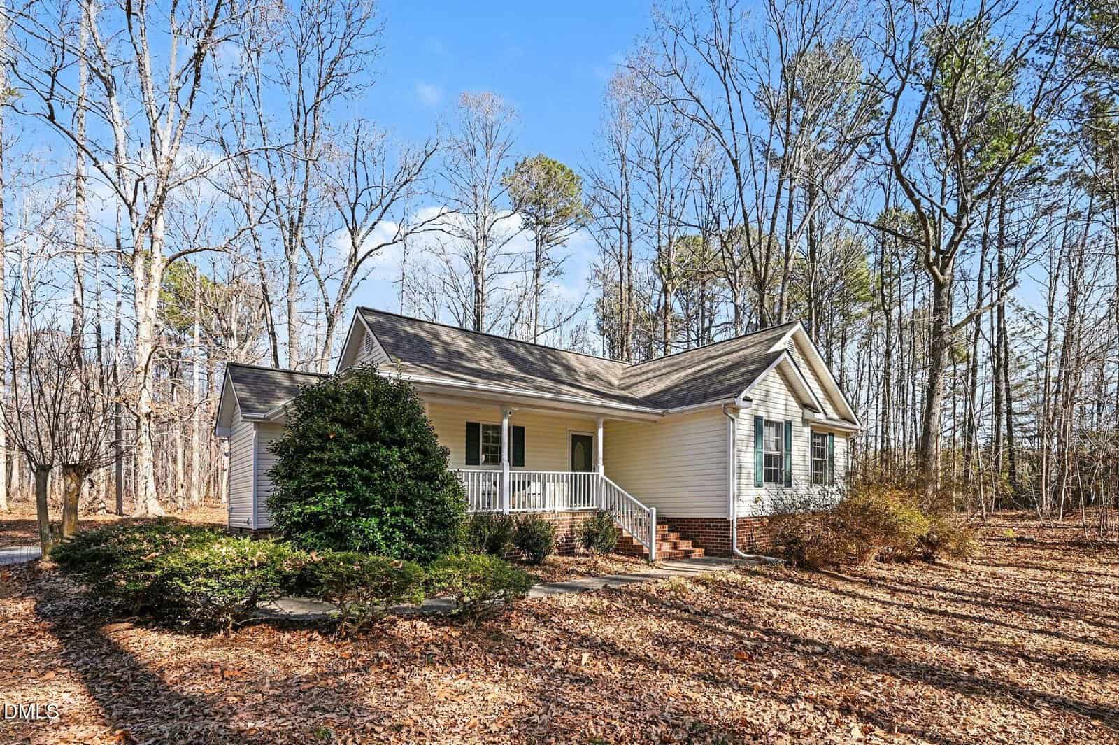 A single-story house with light-colored siding, a front porch with white railings, and steps leading up to the entrance. The home is surrounded by leafless trees and shrubs on a sunny day.