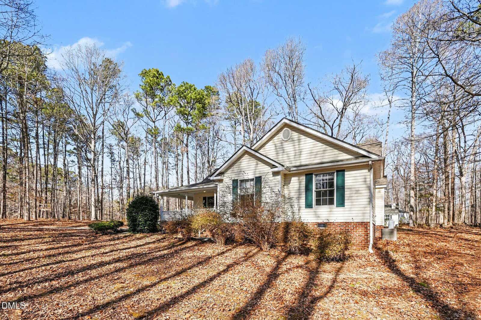 Single-story house with light-colored siding and green shutters, surrounded by leafless trees and fallen leaves. The house has a brick foundation, a small front porch, and is set in a wooded area under a clear sky.