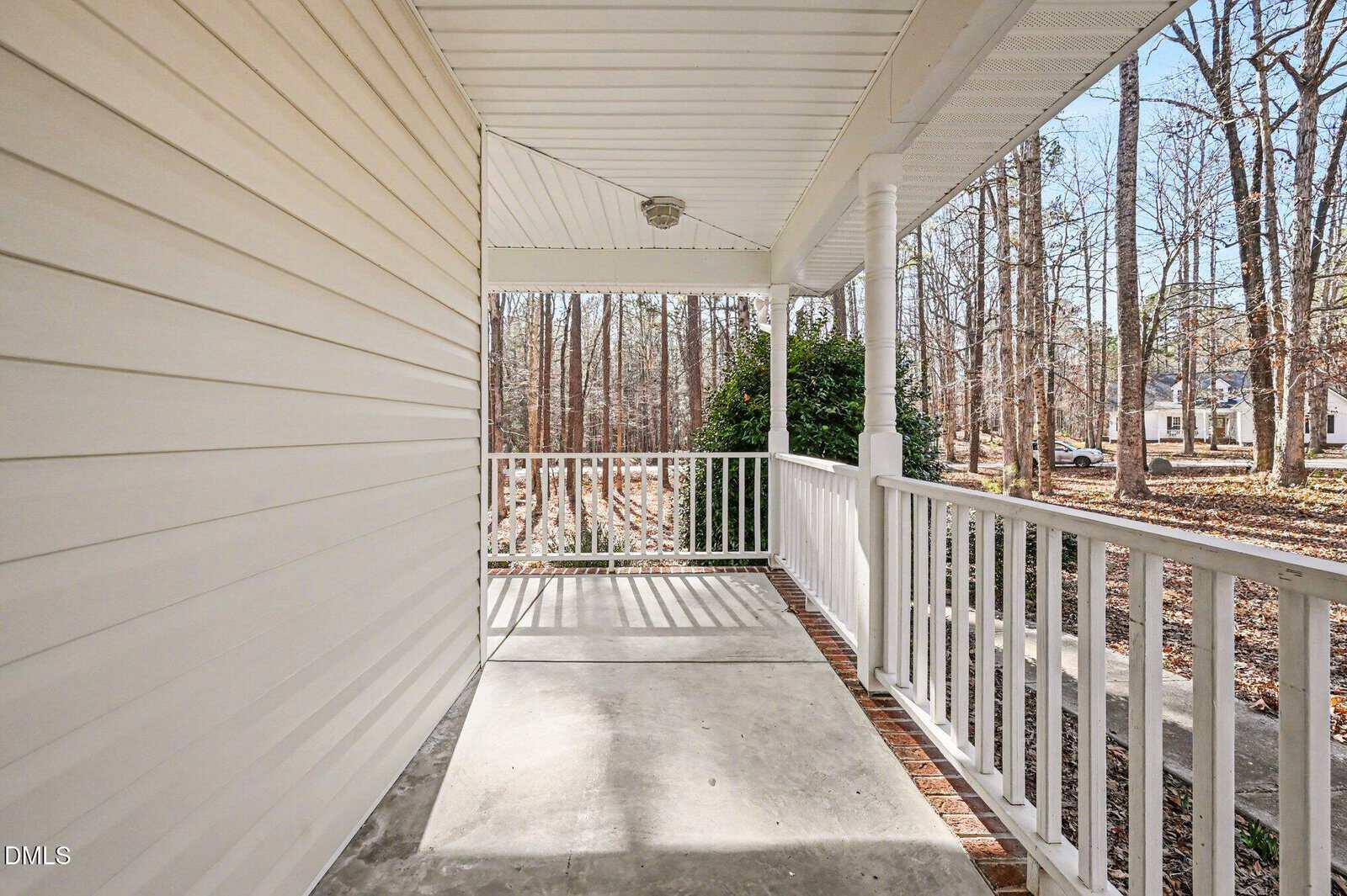 A covered front porch with white railings and columns, concrete floor, and siding. The porch overlooks a yard with trees and a street with houses visible in the background.