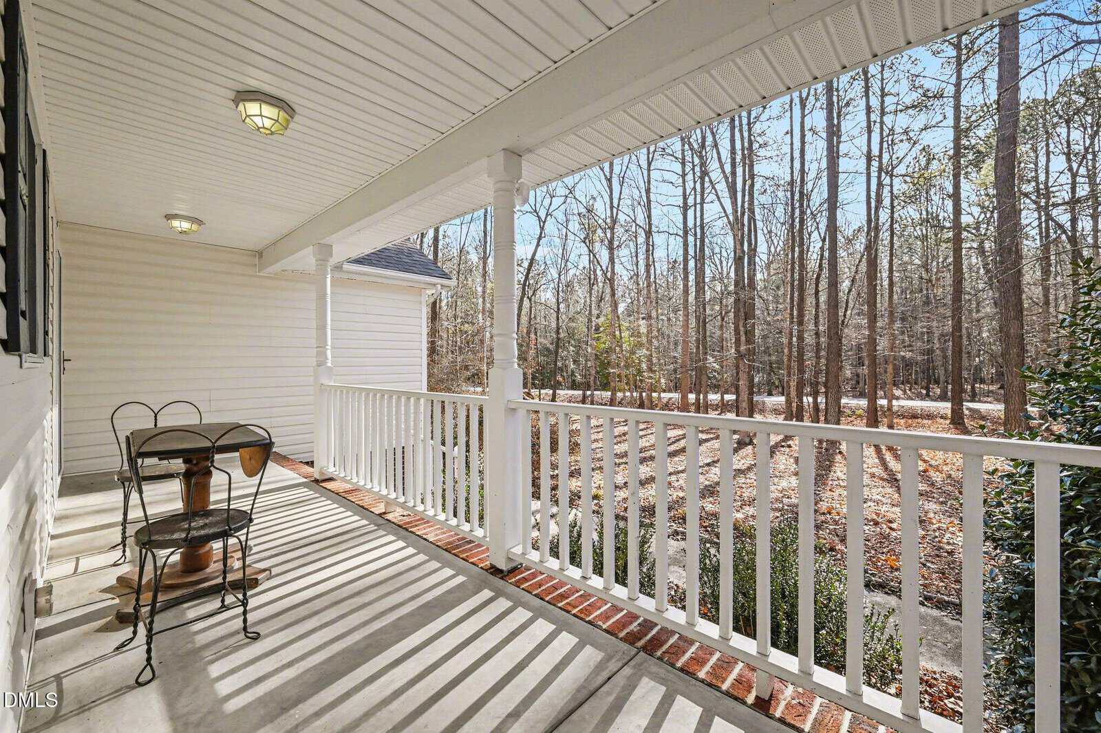 A white porch with a round table and two chairs overlooks a wooded area with leafless trees. Sunlight casts shadows on the floor, creating a calm, inviting outdoor space.