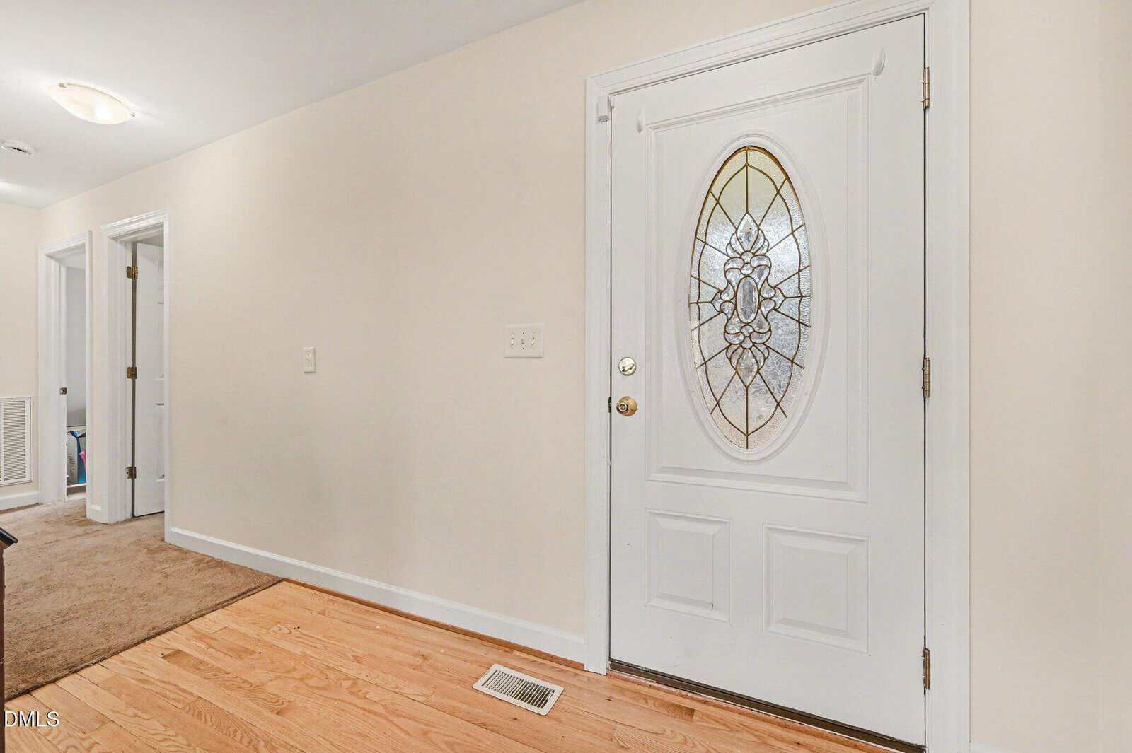 A white front door with an oval glass window and decorative design, set in a hallway with light-colored walls and hardwood flooring. A carpeted area and open doorways are visible in the background.