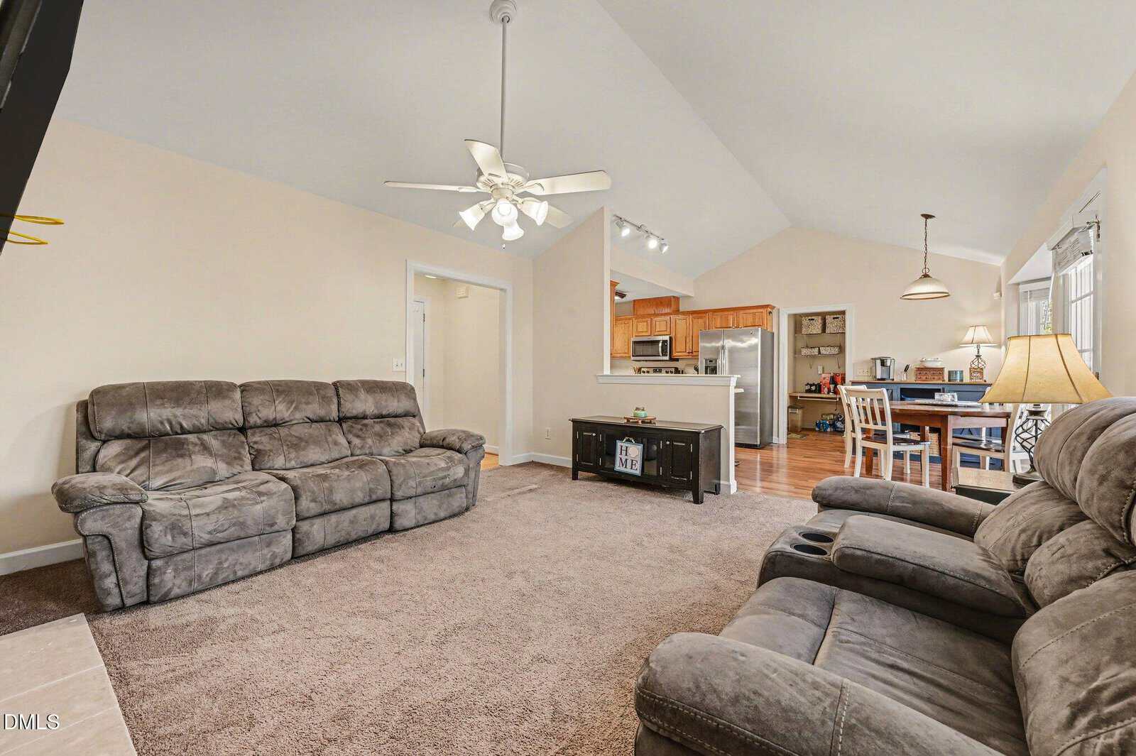 A spacious living room with two gray recliners, carpeted floor, ceiling fan, and view into a kitchen with wooden cabinets and a dining area with a table and chairs. Natural light fills the room from windows.