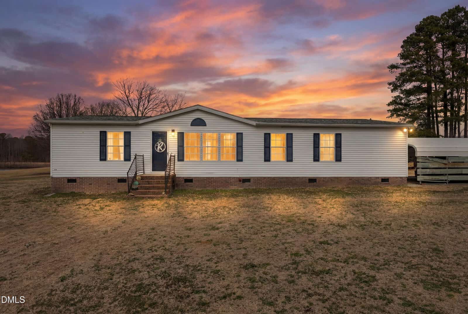 Single-story white manufactured home with blue shutters and brick foundation, lit from inside, sits on a large grassy lot at sunset with a colorful sky and tall trees in the background at 243 Carlyle Road in Zebulon.
