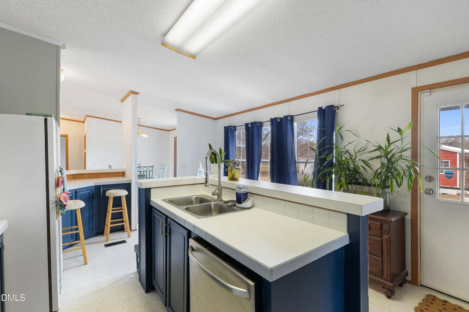 A kitchen with blue cabinets, white countertops, a double sink, and stainless steel dishwasher. Bar stools sit by a counter, and large windows with blue curtains let in natural light. A potted plant is near a glass door.
