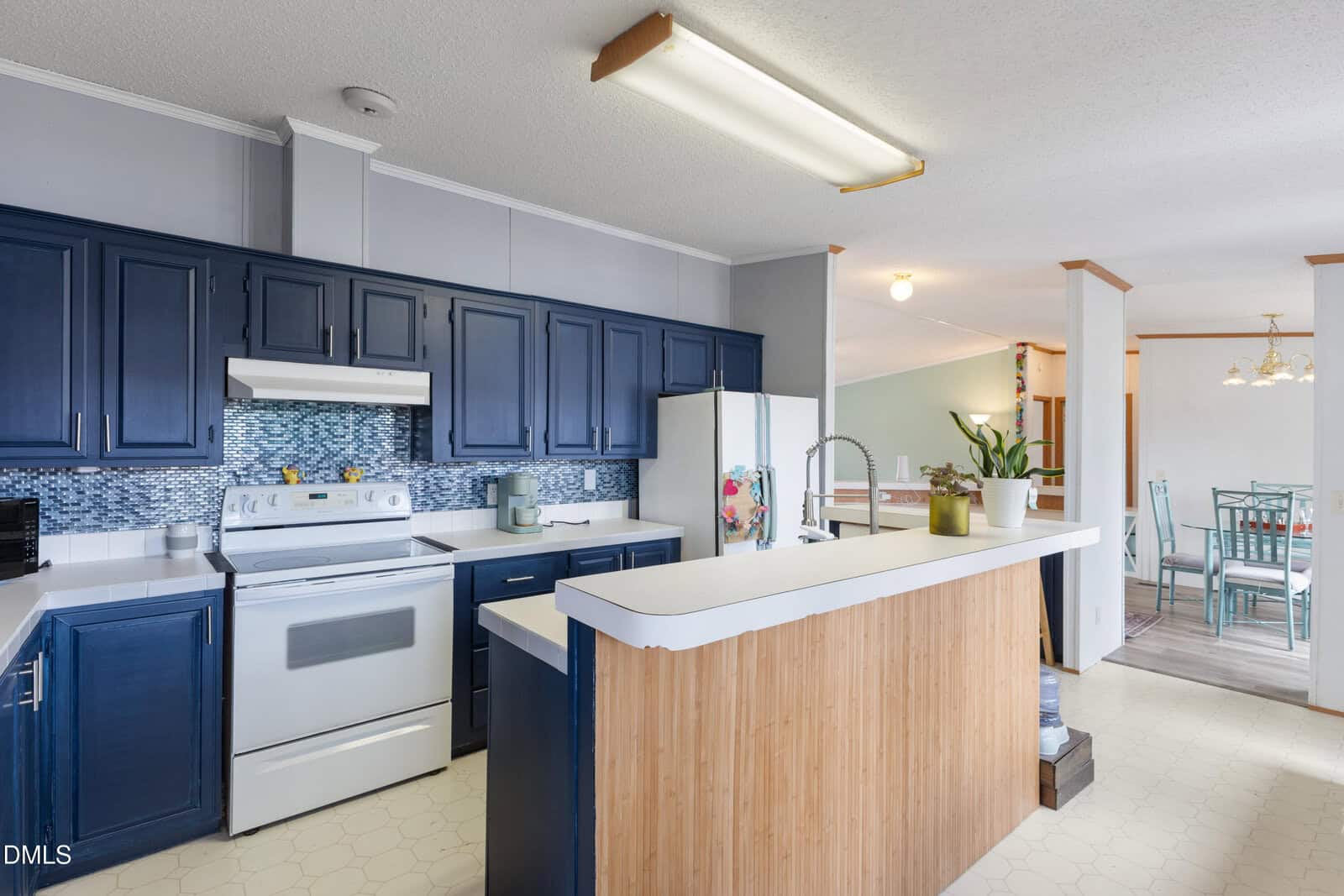 Modern kitchen with blue cabinets, white appliances, a patterned backsplash, and a wood-accented island. Dining area with a glass table and chairs is visible in the background. Ceiling lights illuminate both spaces.