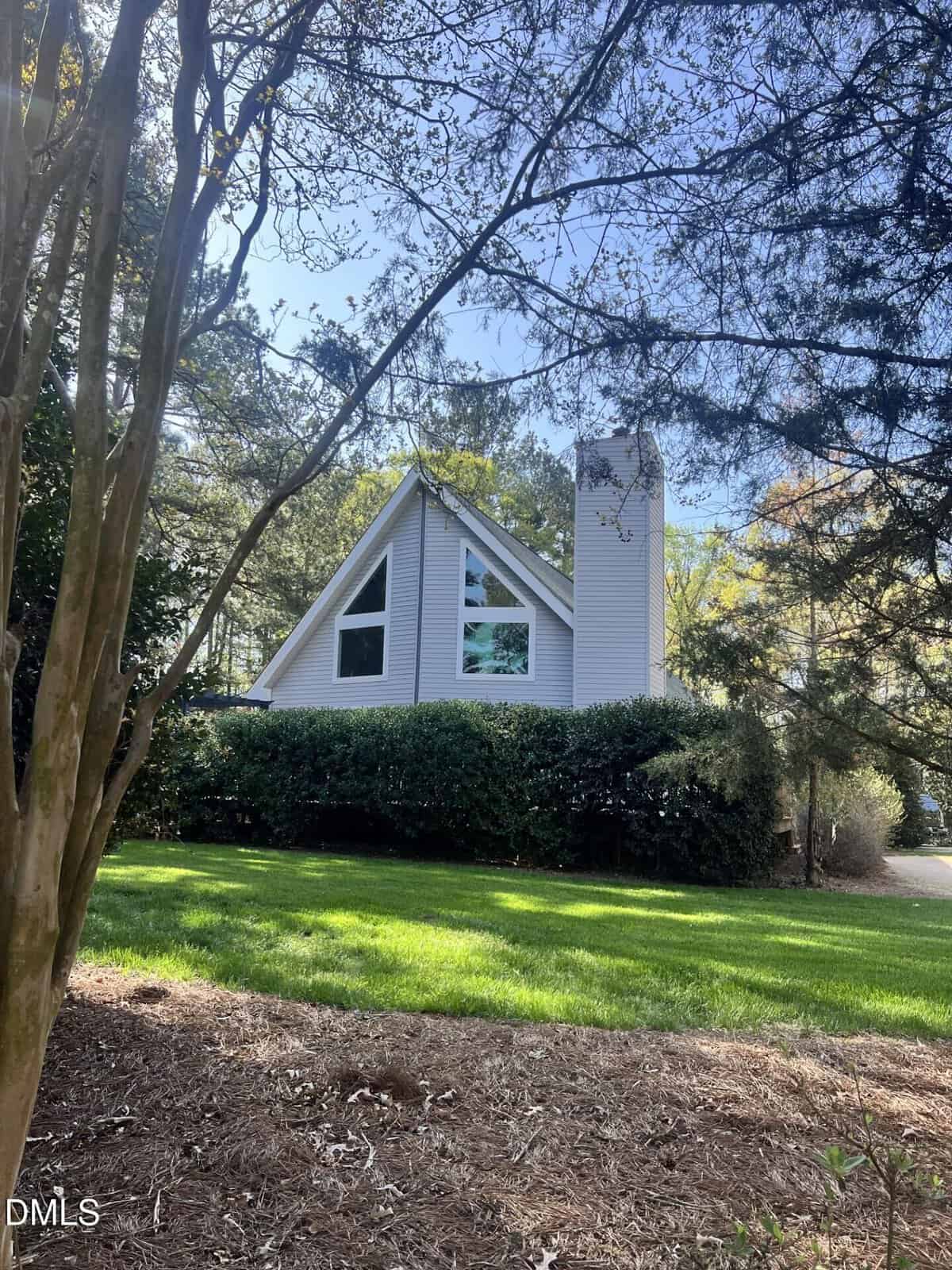 A-frame house with large triangular windows and a tall white chimney, surrounded by green grass, shrubs, and trees on a sunny day.