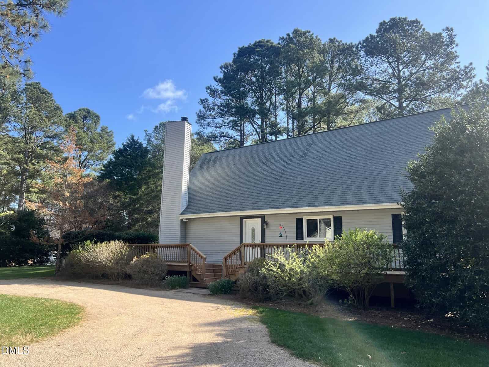 A light gray house with a sloped roof, white trim, and a front porch sits among trees and bushes under a blue sky. A gravel driveway curves in front of the house.