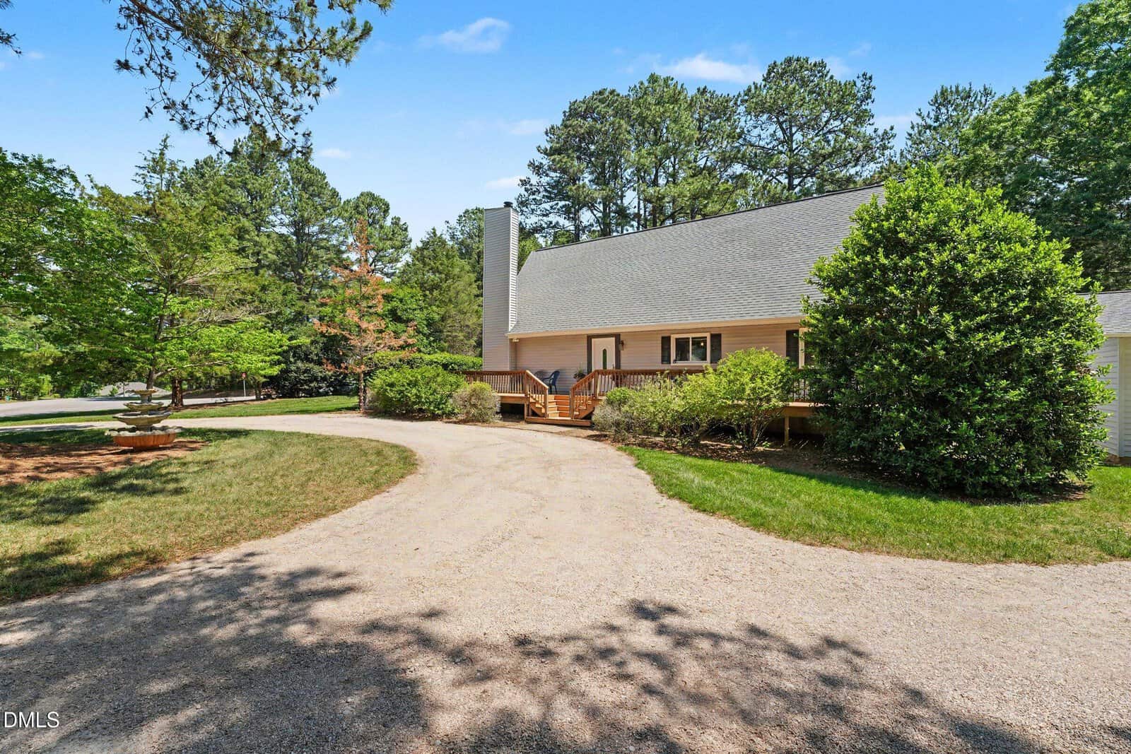 A light-colored house with a steep roof sits next to a circular gravel driveway, surrounded by green trees and bushes under a blue sky.