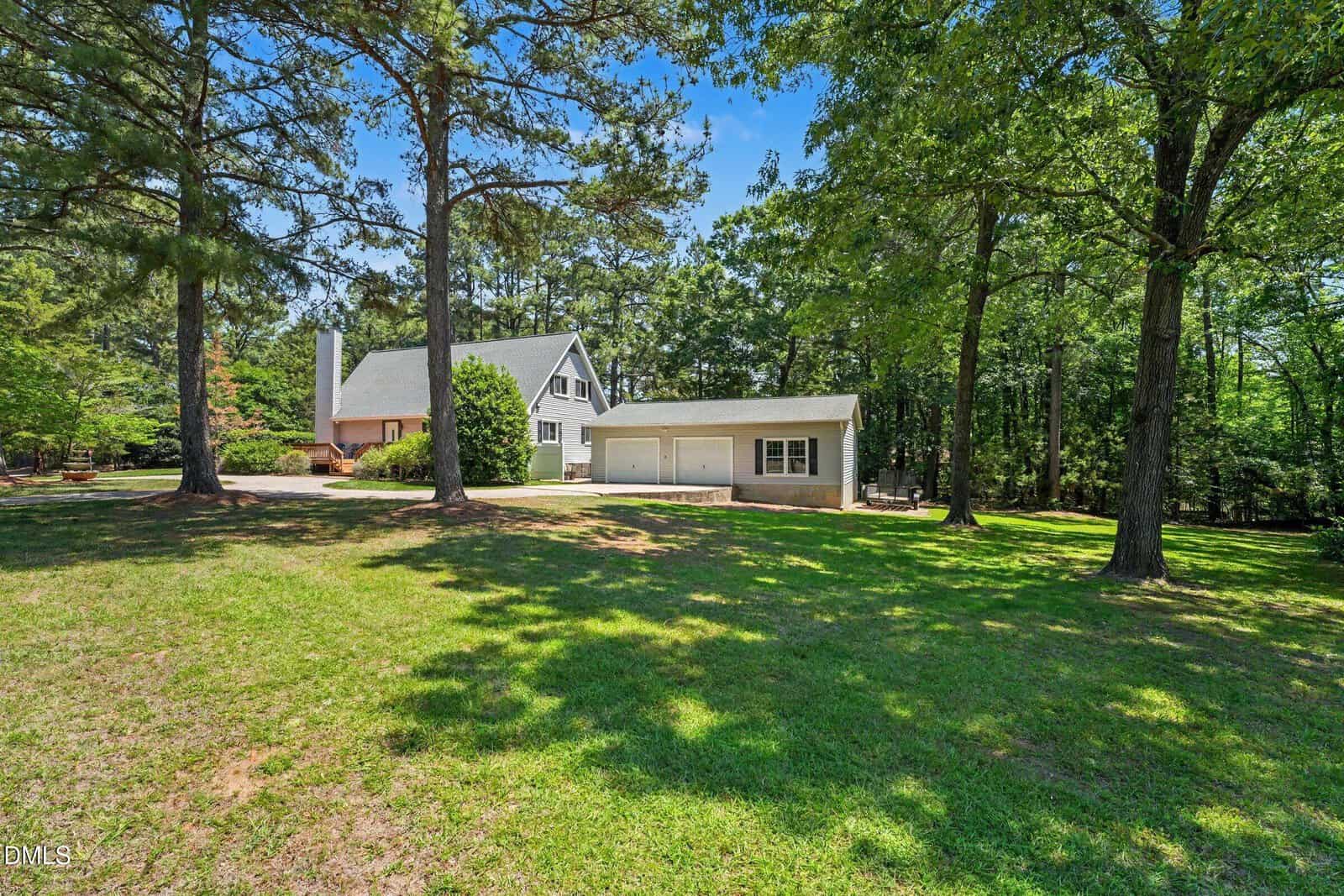 A white, two-story house with a gabled roof and attached garage sits among tall trees on a large, grassy lawn under a blue sky. Shadows from the trees cover parts of the yard.