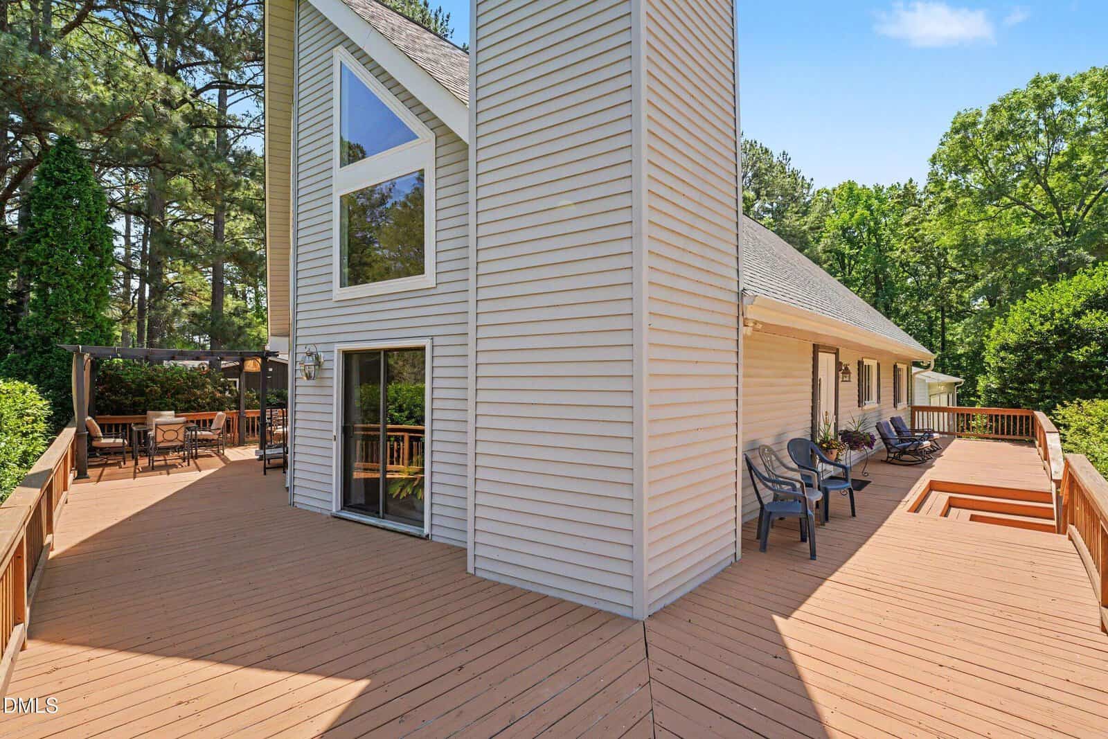 A spacious wooden deck wraps around a modern house with large windows and beige siding. Patio chairs and a covered dining area are visible, surrounded by trees under a clear blue sky.