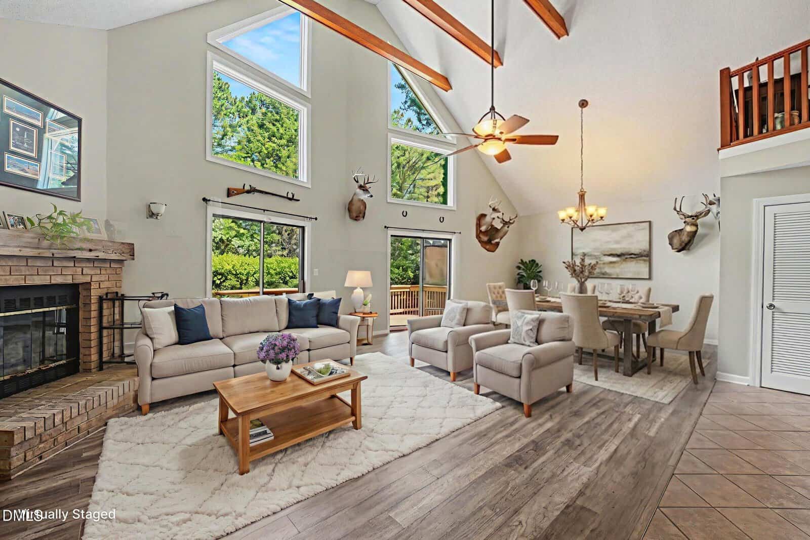 A spacious living and dining area at 110 Mohican Drive in Louisburg features vaulted ceilings, exposed wooden beams, large windows, a fireplace, beige sofas, armchairs, a wooden coffee table, dining set, and mounted deer heads on the walls.