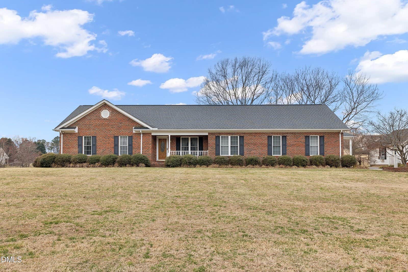 A single-story brick house with dark roof shingles, white trim, and shutters is nestled at 102 Justin Lane in Louisburg. There is a front porch with columns and bushes lining the house, which sits on a large grassy lawn dotted with a few leafless trees.