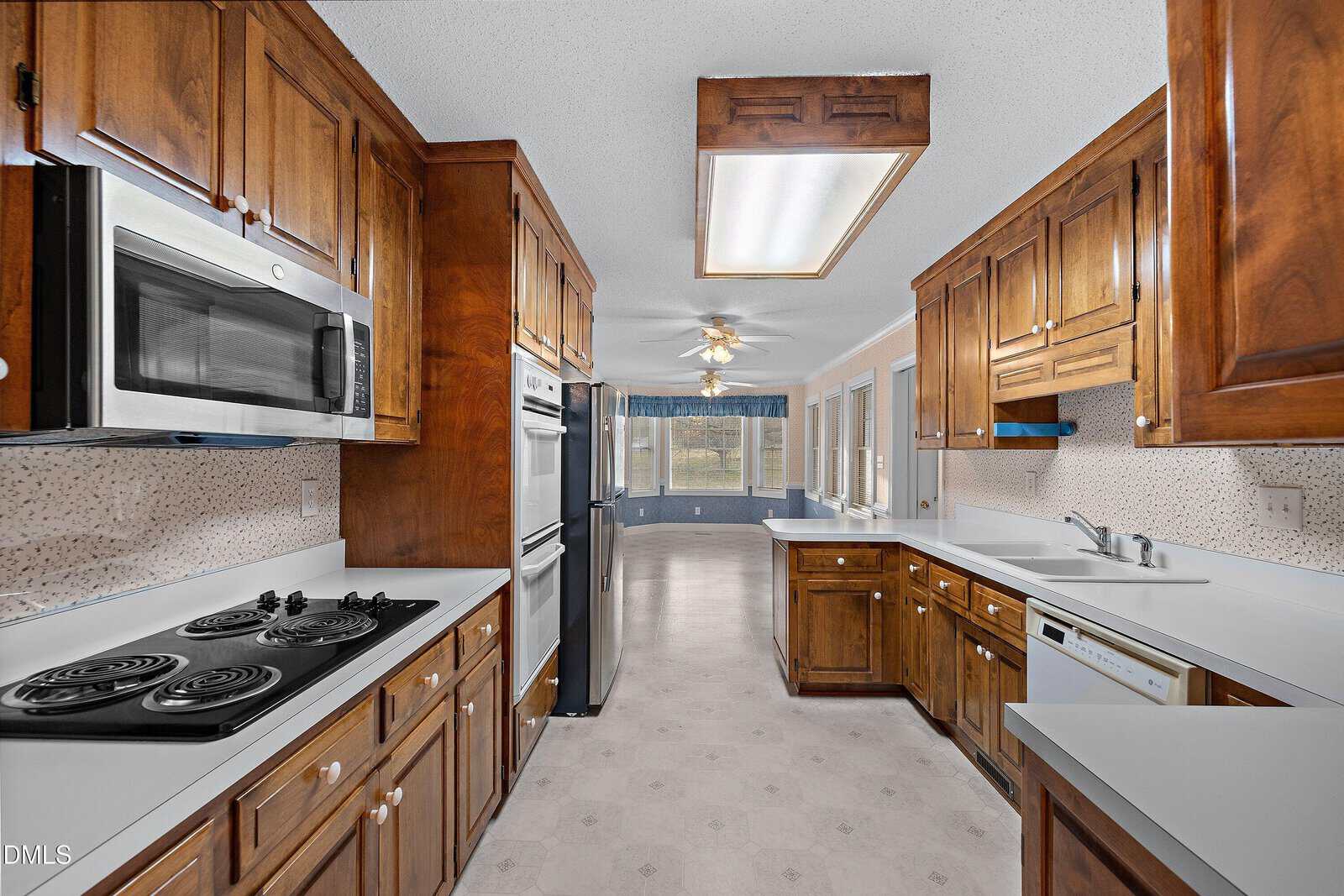 Galley-style kitchen with wooden cabinets, electric stovetop, built-in microwave, stainless steel fridge, dishwasher, dual sinks, ceiling lights, and view into a dining area with large windows and ceiling fans.