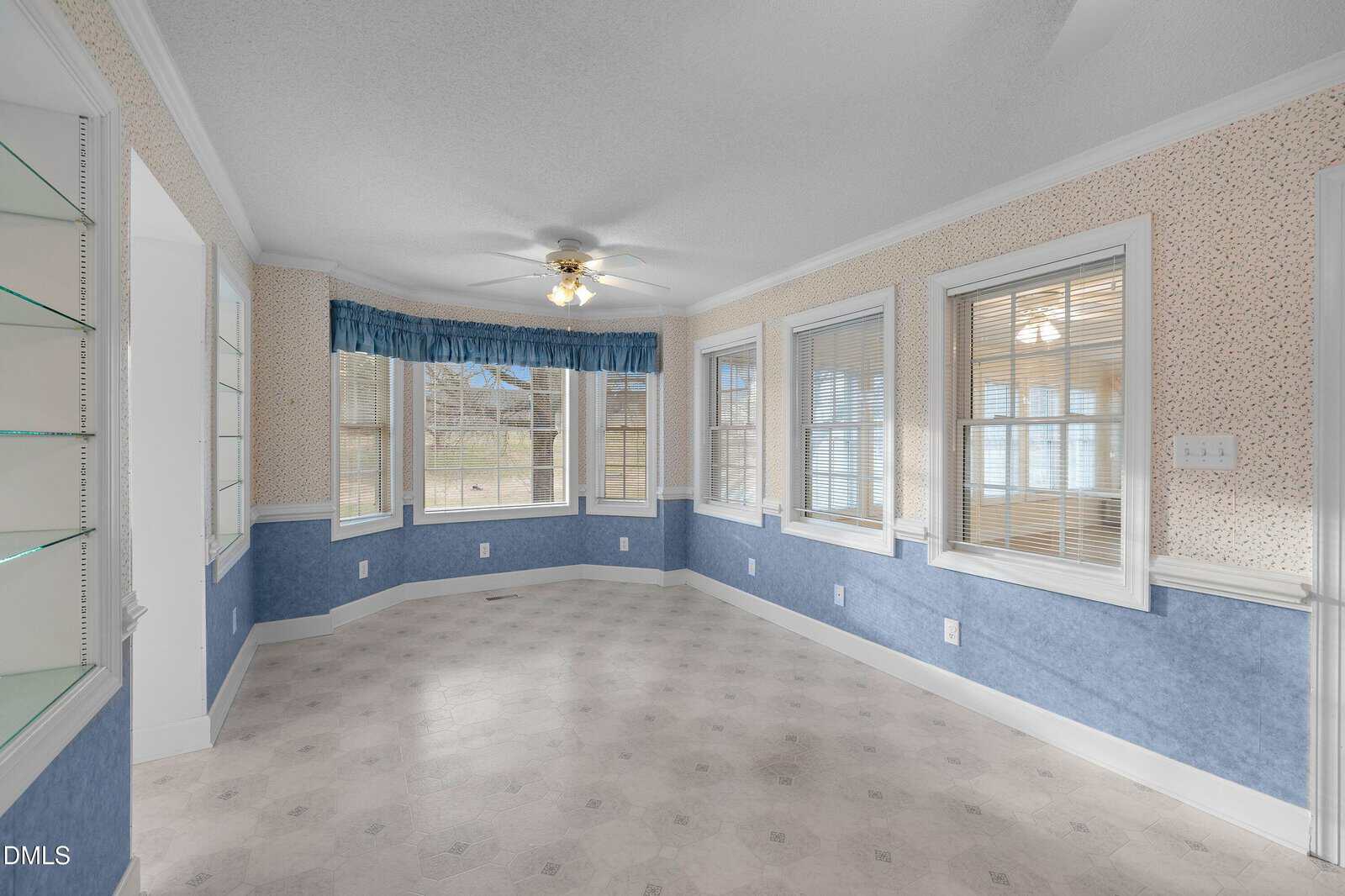 A spacious sunroom with blue and cream wallpaper, large windows on three walls, white trim, tile flooring, and a ceiling fan with a light fixture. Built-in shelves are visible on the left side of the image.