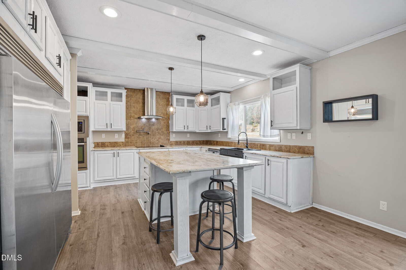 Modern kitchen with white cabinets, stainless steel appliances, a central island with two black stools, pendant lights, wood-style flooring, and a window above the sink. Walls are painted beige and backsplash is tan tile.