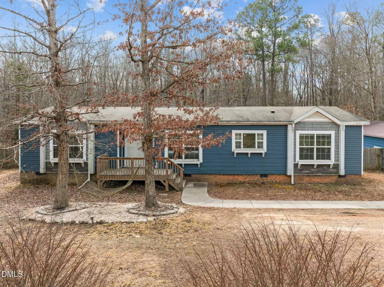 A blue single-story manufactured home with white trim and a small front porch sits among leafless trees at 3200 Granville Ridge Road in Wake Forest, with a curved driveway in front and a wooded area in the background.