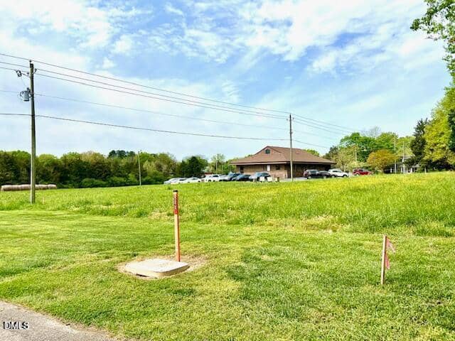 A grassy field at 0 College Street, Oxford, features two red marker stakes in the foreground, power lines overhead, and a single-story building with multiple parked cars in the background under a partly cloudy sky.