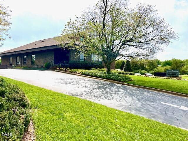 A single-story brick office building sits behind a neatly landscaped lawn with a large tree in front. A paved driveway leads to the entrance, and a sign is visible near the edge of the street.