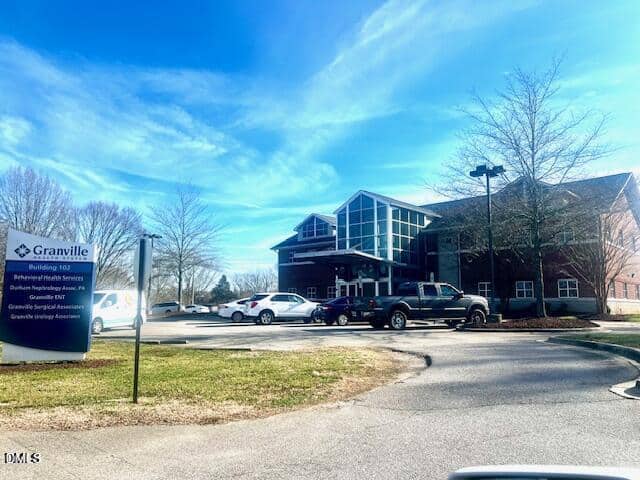 A modern brick medical building with large windows and a parking lot in front. A sign reads “Granville” with several healthcare services listed. Several cars are parked, and leafless trees surround the area under a blue sky.