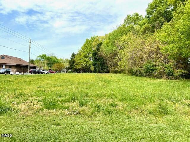 A grassy, open lot bordered by trees under a partly cloudy sky. A building, parked cars, and utility poles are visible on the left side near the edge of the image.