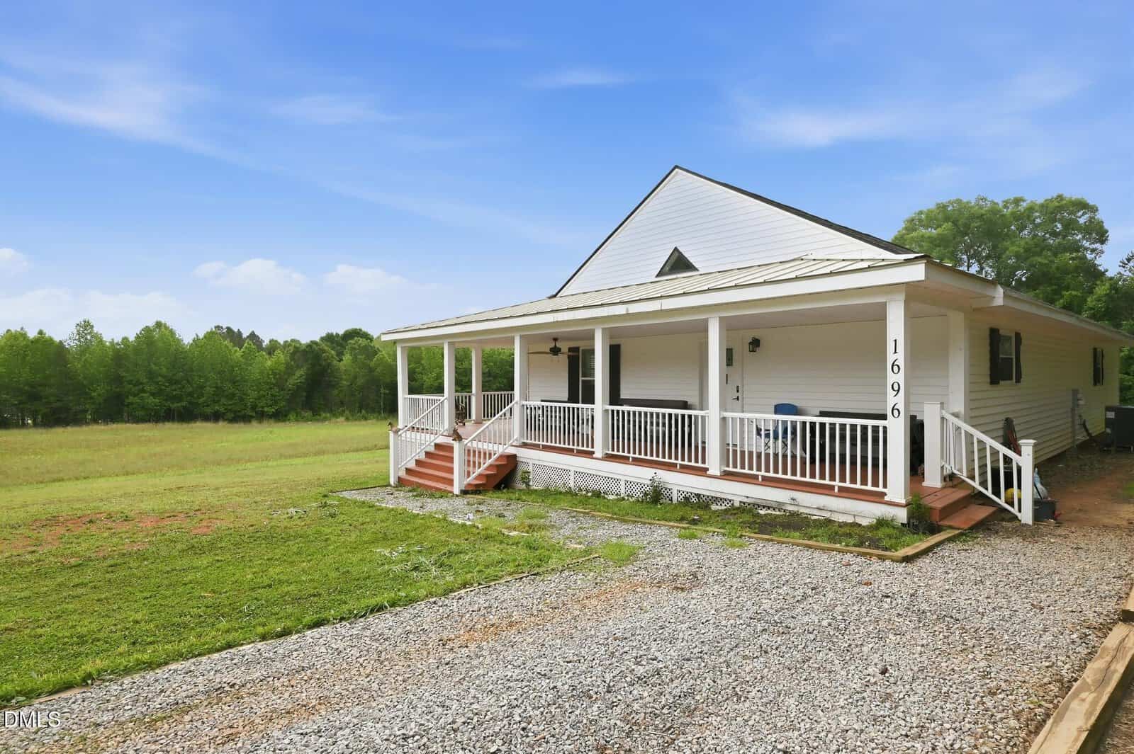 A white single-story house with a large front porch, steps, and railing, sits next to a gravel driveway and open grassy yard, with trees and a blue sky in the background.