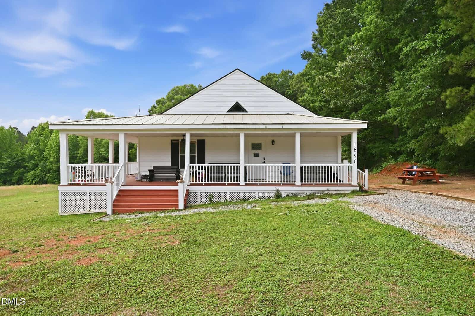 A white house with a wraparound porch and red steps, set on a grassy lawn with trees in the background. A gravel path leads to the porch, and a red picnic table sits to the right near the edge of the woods.