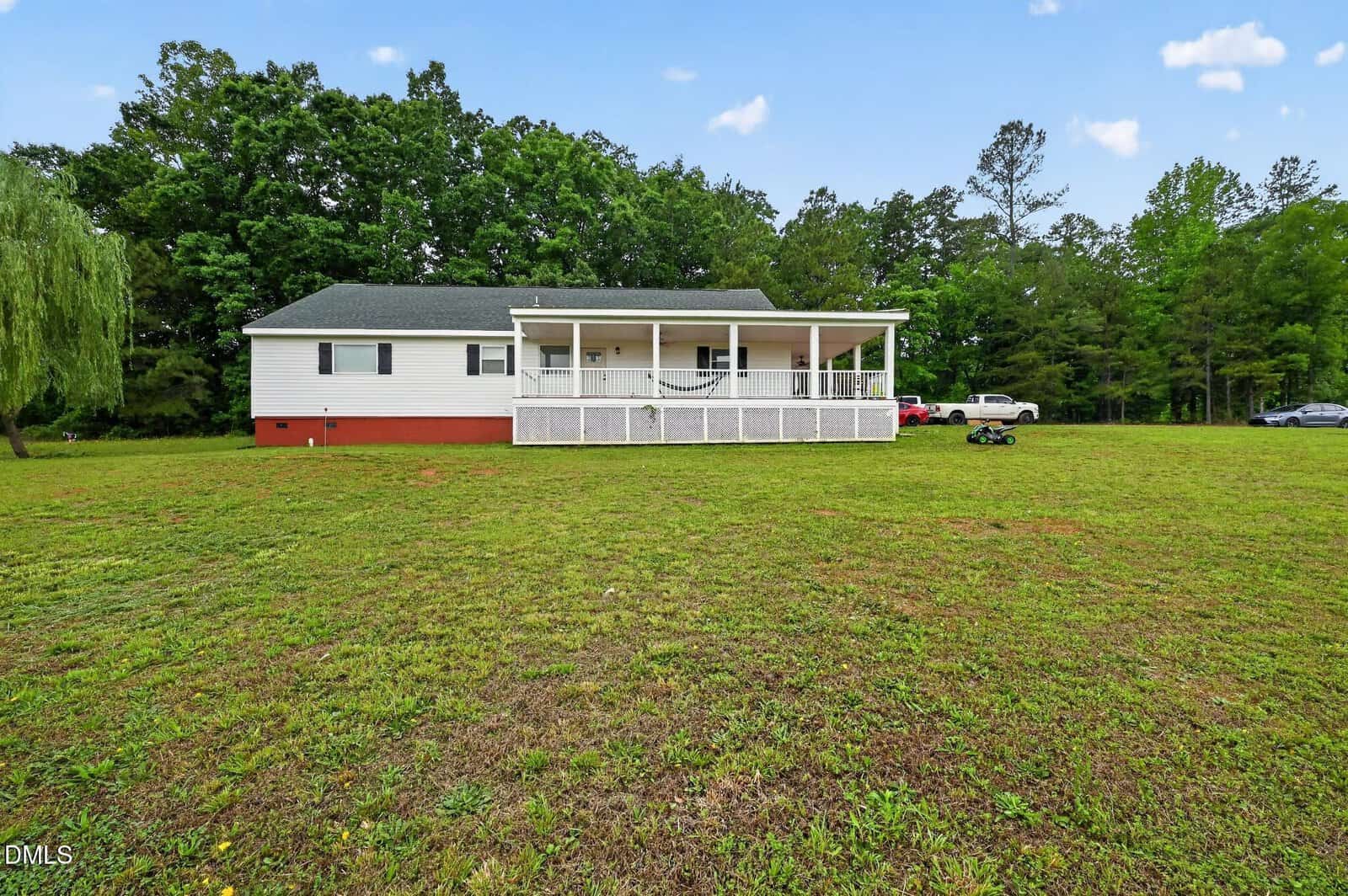 A single-story house with a large covered front porch and white railing sits on a spacious grassy yard, surrounded by trees under a blue sky with scattered clouds.