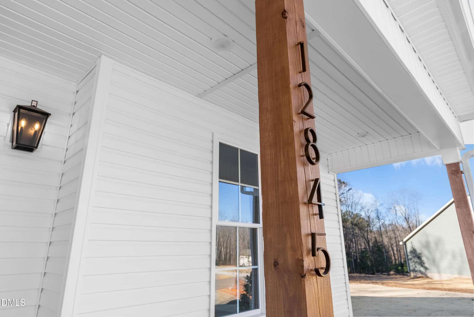 A wooden porch post displays house numbers 12845 vertically. The white house exterior features horizontal siding, a window, a covered entryway ceiling, and a lit black wall lantern. Trees and another house are visible in the background.