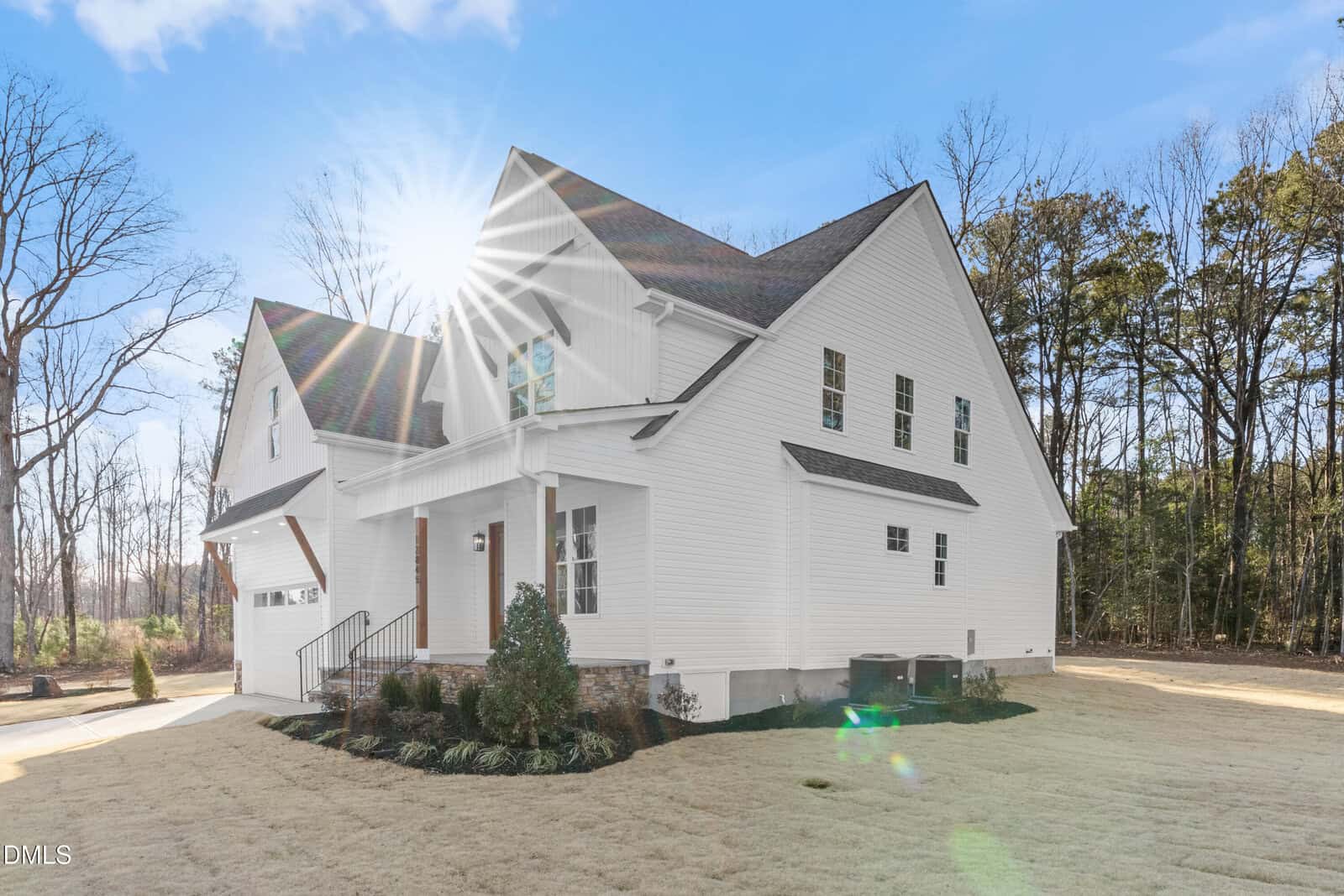 A white two-story house with a front porch, attached garage, and manicured lawn under a clear sky. Sunlight creates a lens flare in the corner; trees line the background.