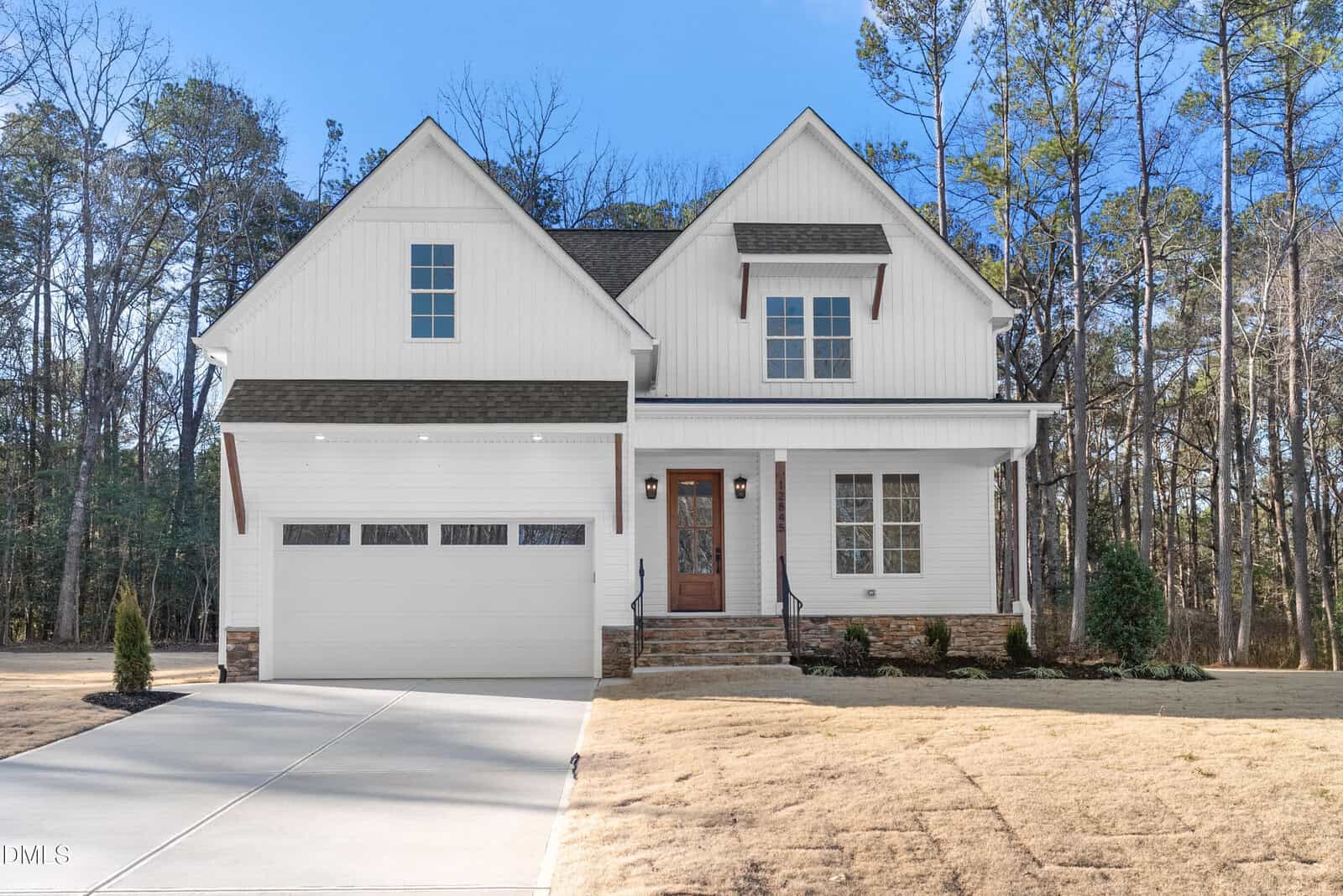 A two-story white house with a front porch, brown wooden door, double garage, and gable roof, set against a backdrop of trees with a concrete driveway and minimal landscaping in the yard, located at 12845 Mallard Rock Road in Zebulon.