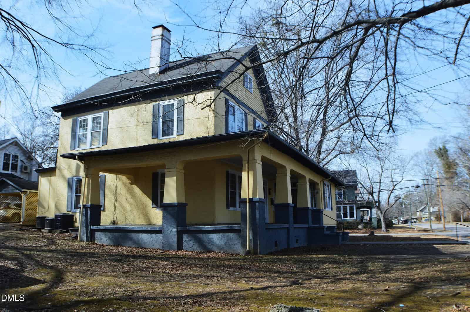 A two-story yellow house with a dark roof, white-trimmed windows, and a covered wraparound porch sits on a bare yard with scattered leaves, surrounded by leafless trees on a clear day. A residential street runs alongside.