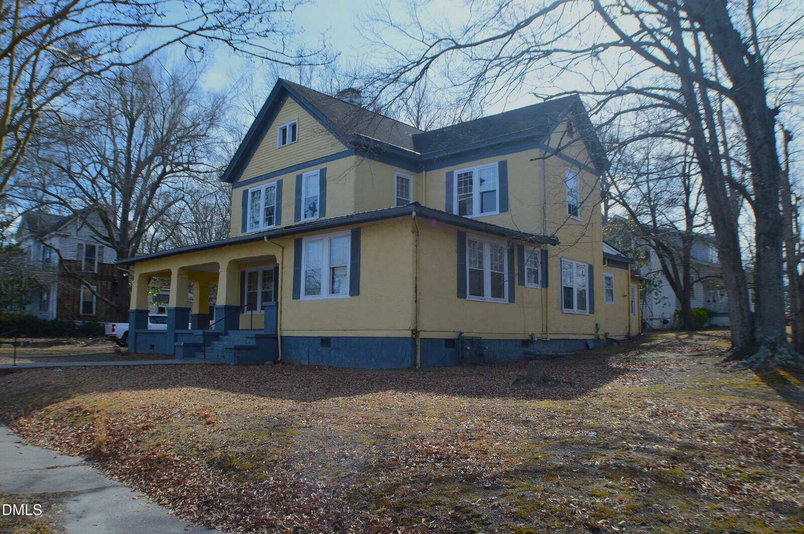 A two-story yellow house with blue trim sits on a corner lot. The house has multiple windows, a covered porch, and leaf-covered ground. Leafless trees and other homes are visible in the background.