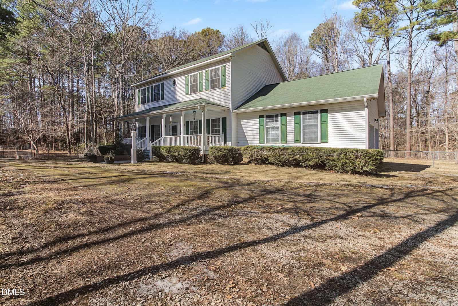 Two-story white house with green shutters and a green roof, surrounded by leafless trees and bushes, with a covered front porch and large yard under clear blue sky.