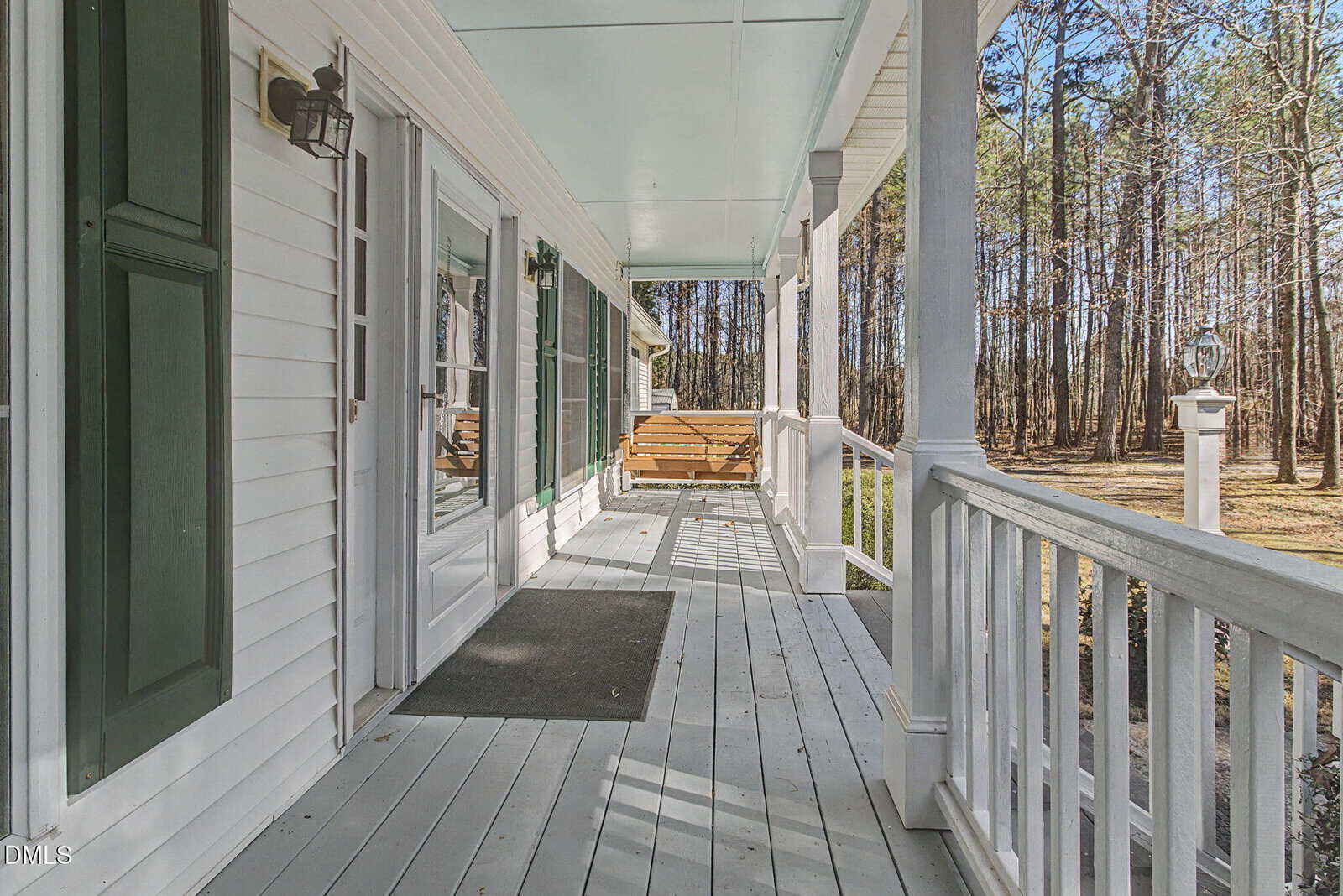 A white wooden porch with green shutters, a bench, light fixture, and railing. The porch overlooks a yard with grass and tall trees in the background under a clear sky.