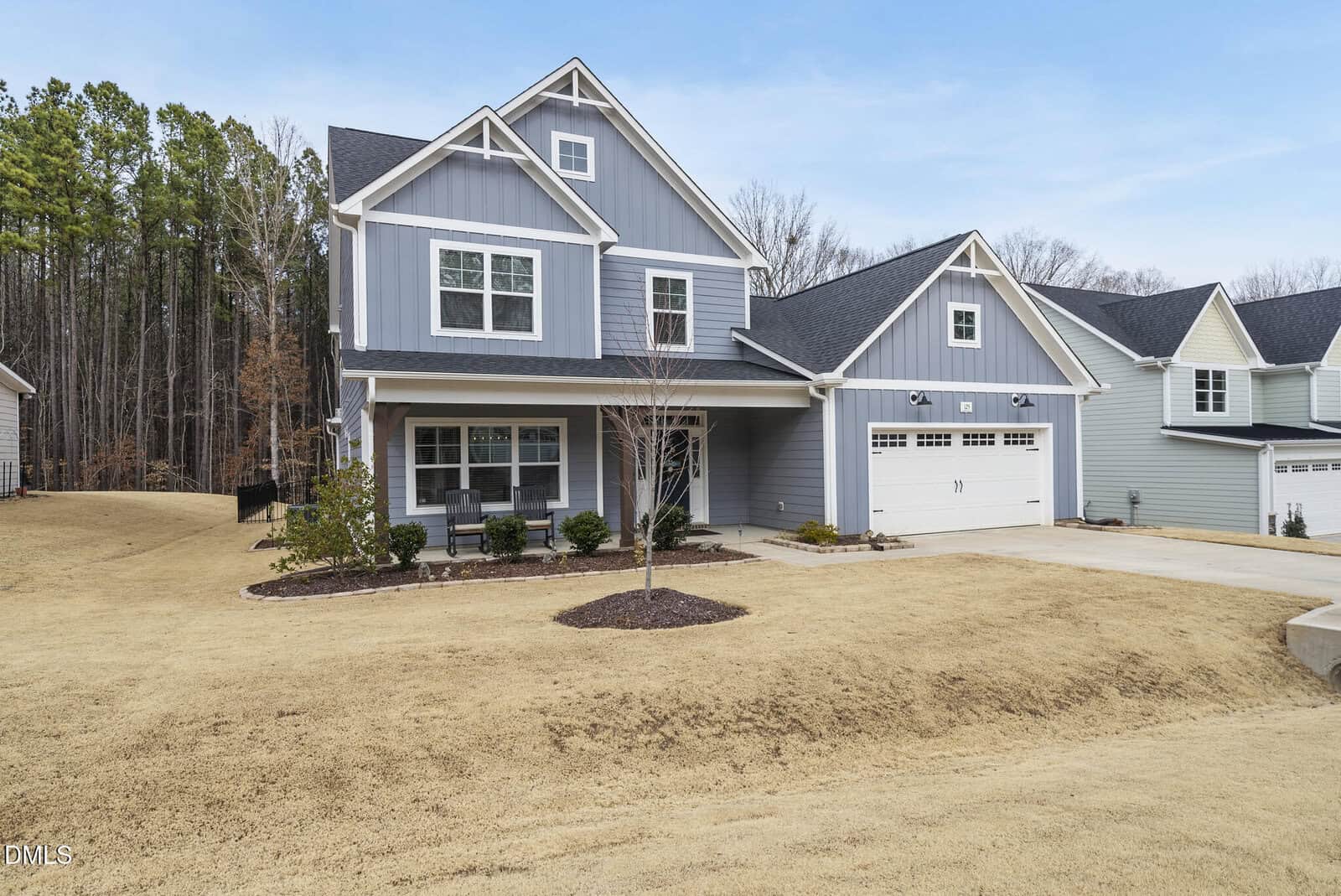 Two-story gray house with white trim, double garage, covered front porch, and a small tree in the front yard. The lawn is dry, and trees line the background under a clear blue sky.