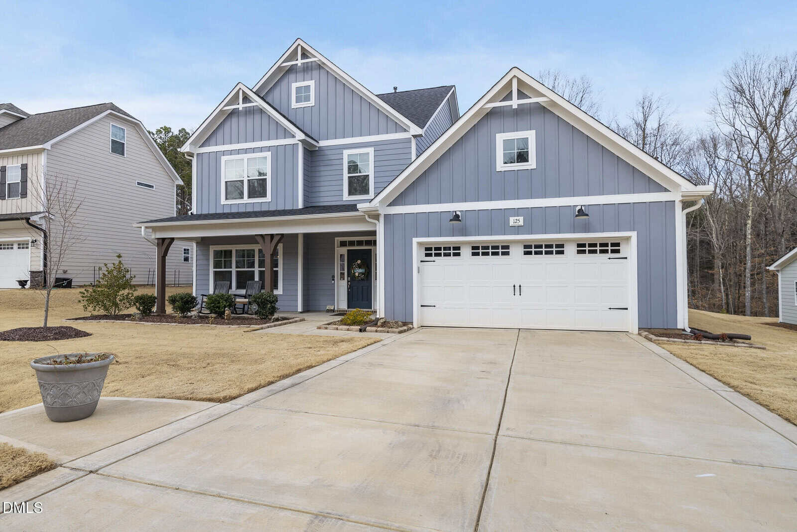 A two-story blue house with white trim, a front porch, and a double garage, sits on a concrete driveway with a manicured lawn and landscaped bushes, surrounded by neighboring homes and trees.
