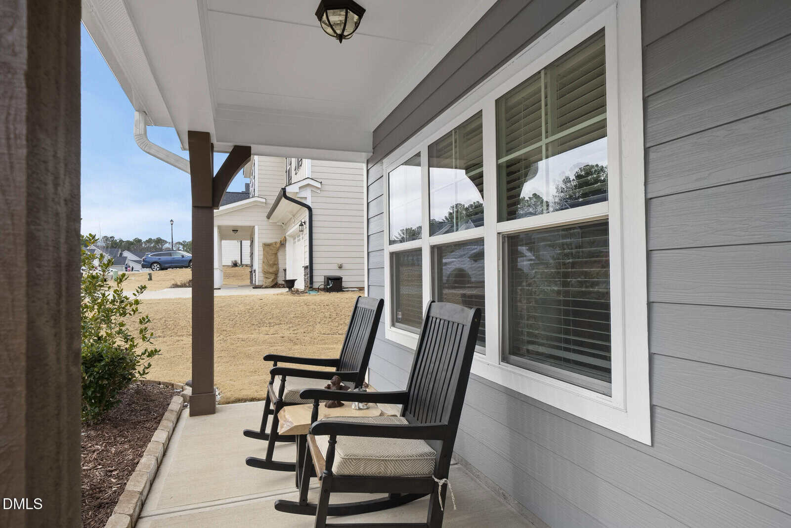 Two black rocking chairs with beige cushions sit on a covered front porch of a gray house with white trim, facing a yard with mulch and small bushes. A light fixture hangs from the porch ceiling.