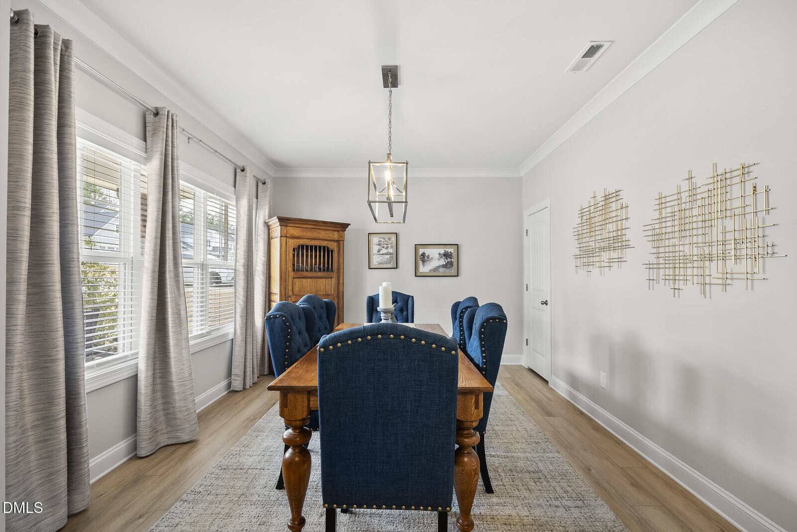 Elegant dining room with a wooden table, six blue upholstered chairs, large windows with gray curtains, light walls, modern chandelier, and geometric wall art. Natural light fills the space.
