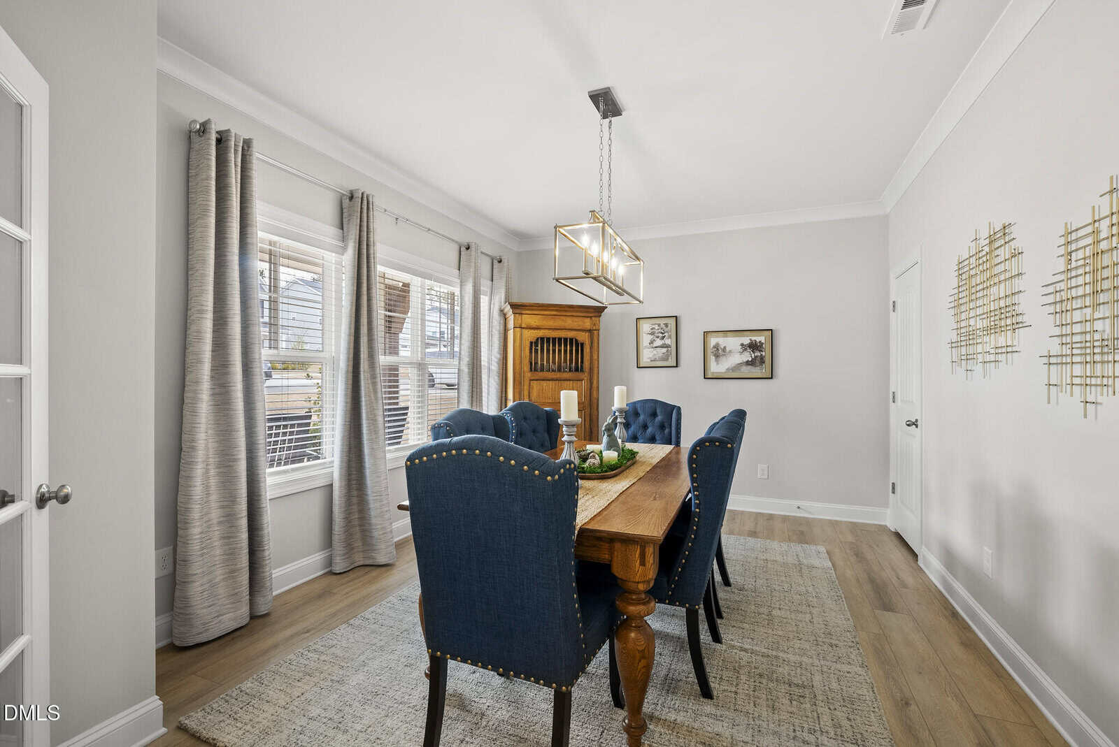 A modern dining room with a wooden table, six blue upholstered chairs, large windows with gray curtains, wall art, and a light fixture above the table. Light wood flooring and neutral walls create a bright, airy feel.