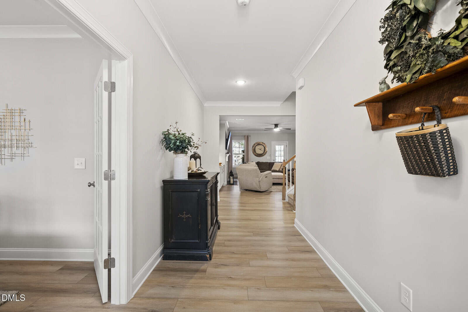 A hallway with light wood floors, beige walls, a black console table with decor, and a shelf with a basket and wreath. It leads to a living area with sofas and a ceiling fan.