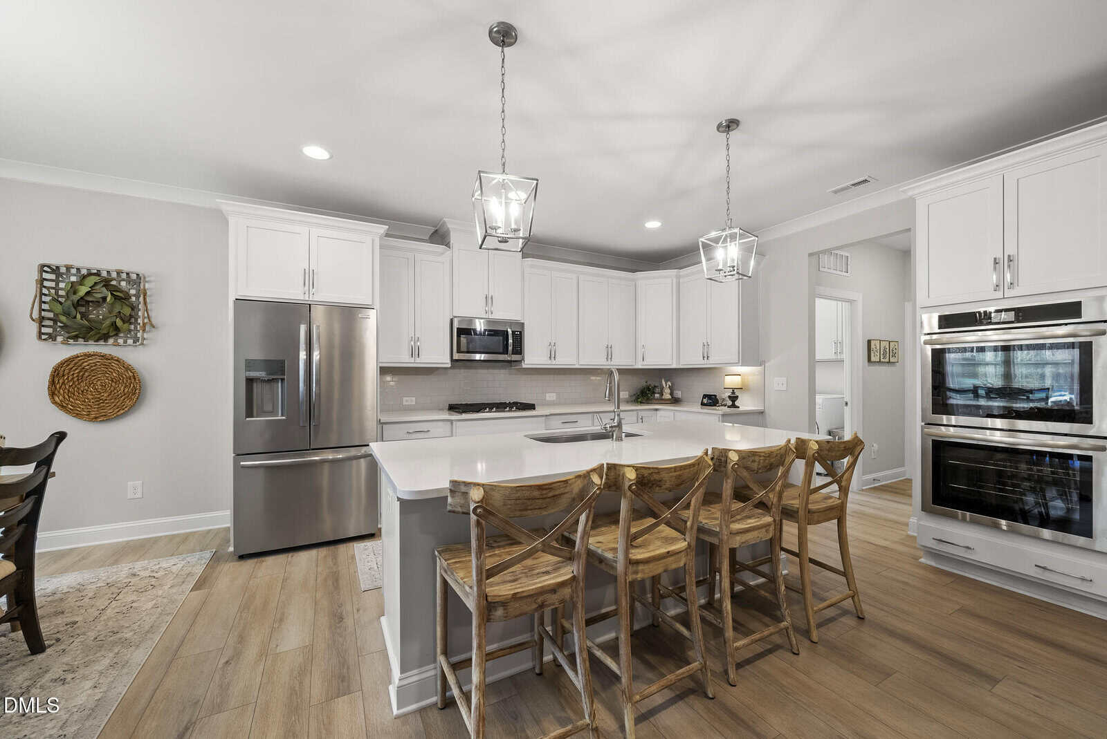 Modern kitchen with white cabinets, stainless steel appliances, a large island with a white countertop, and four wooden barstools. Pendant lights hang above the island, and wood flooring is throughout the space.