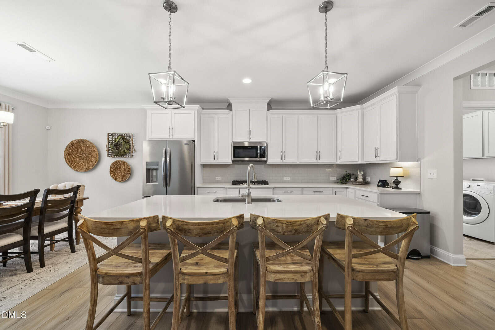 Modern kitchen with white cabinets, stainless steel appliances, large island with four wooden barstools, pendant lights above, and a dining area to the left. A laundry room is visible on the right.