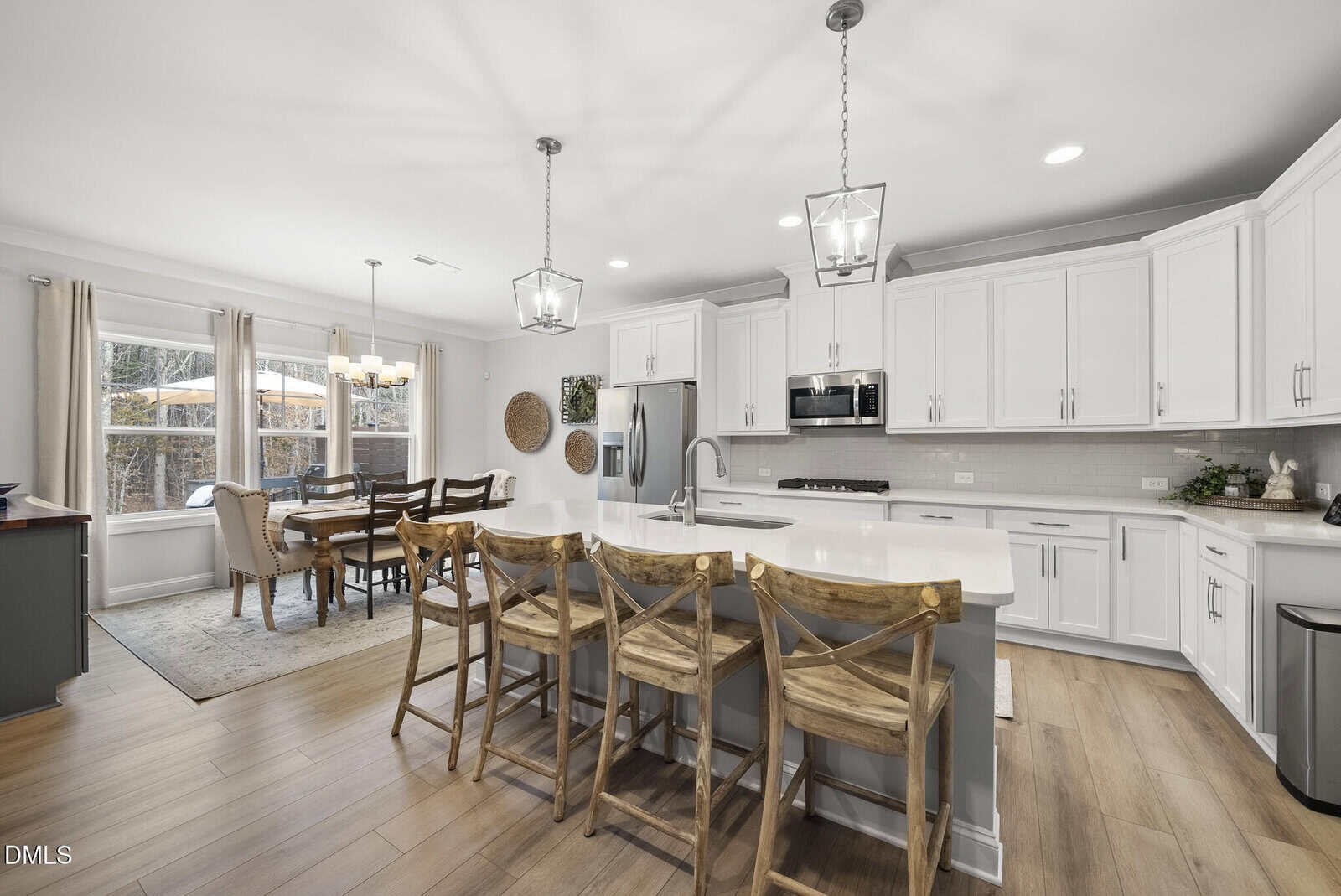 Modern kitchen with white cabinets, stainless steel appliances, a large island with a sink, and three wooden barstools. Adjacent dining area features a wooden table and six chairs near large windows with beige curtains.