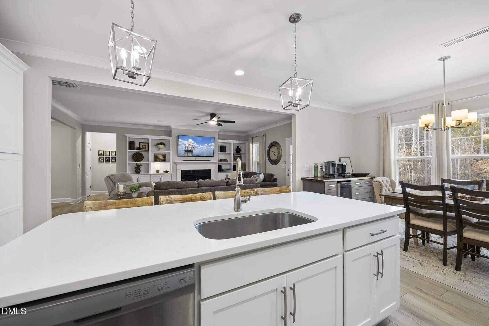 Modern open-concept kitchen with a white island and sink, overlooking a living room with a large sectional sofa, TV, and built-in shelves. A dining area with a wooden table is on the right near large windows.