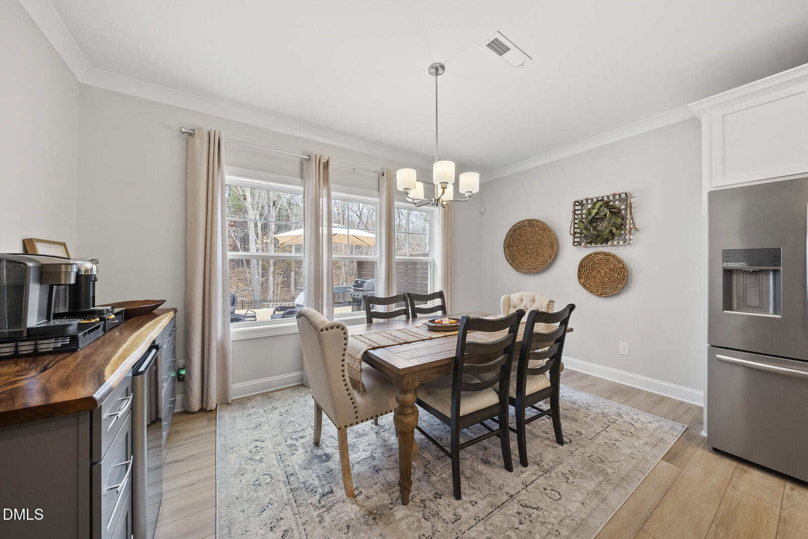 A bright dining area with a wooden table, four chairs, a tufted bench, and a chandelier. Large window with beige curtains lets in natural light. Three woven wall decorations hang beside a stainless steel refrigerator.