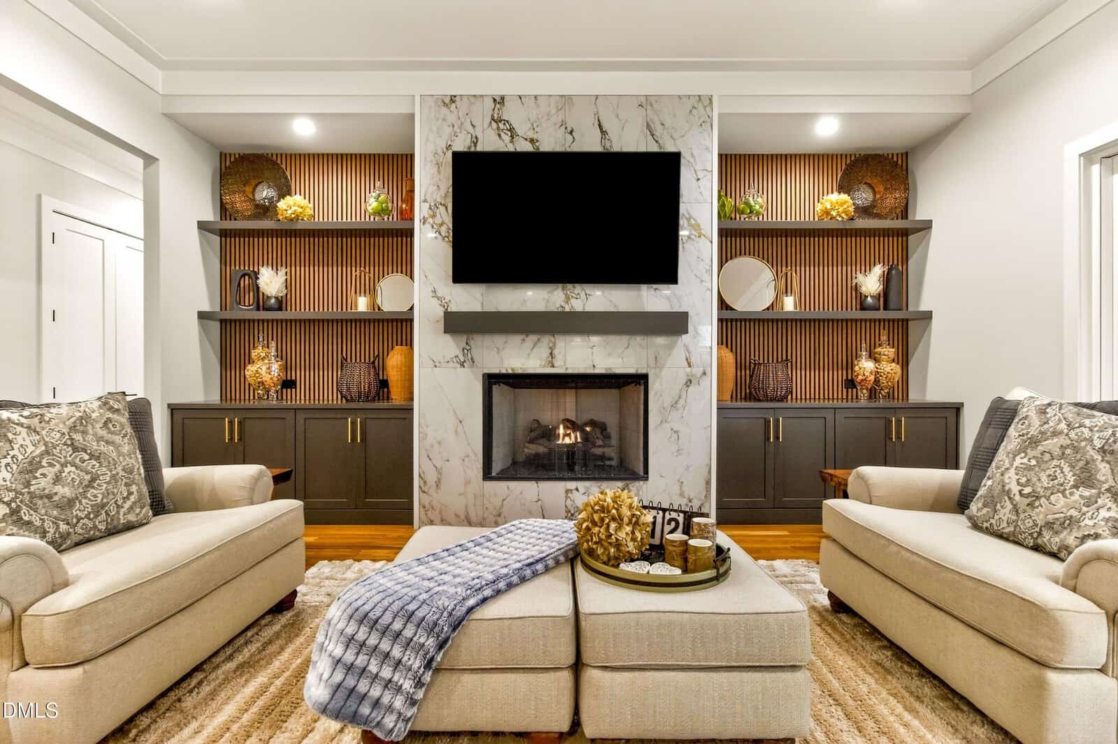Modern living room with two beige armchairs, a marble fireplace, and a mounted TV. Built-in shelves display decor, and a cozy rug and throw blanket add warmth. Neutral tones create a welcoming, elegant atmosphere.