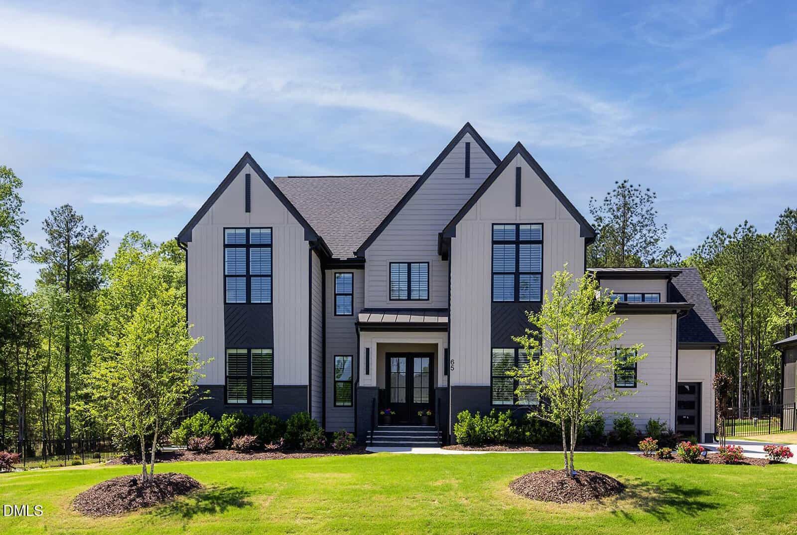 A modern two-story house with light vertical siding, dark trim, large windows, and gabled roofs, surrounded by a green lawn, small trees, and landscaping, with a wooded area in the background under a blue sky.