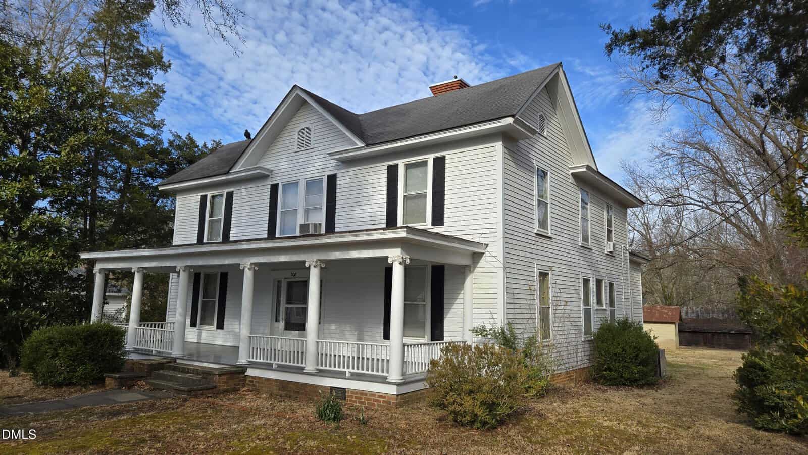 Two-story white wooden house with black shutters, a covered front porch with columns, and a gable roof. Nestled on 308 Sunset Avenue in Oxford, it is surrounded by grass, bushes, trees, and a small outbuilding in the background under a partly cloudy sky.