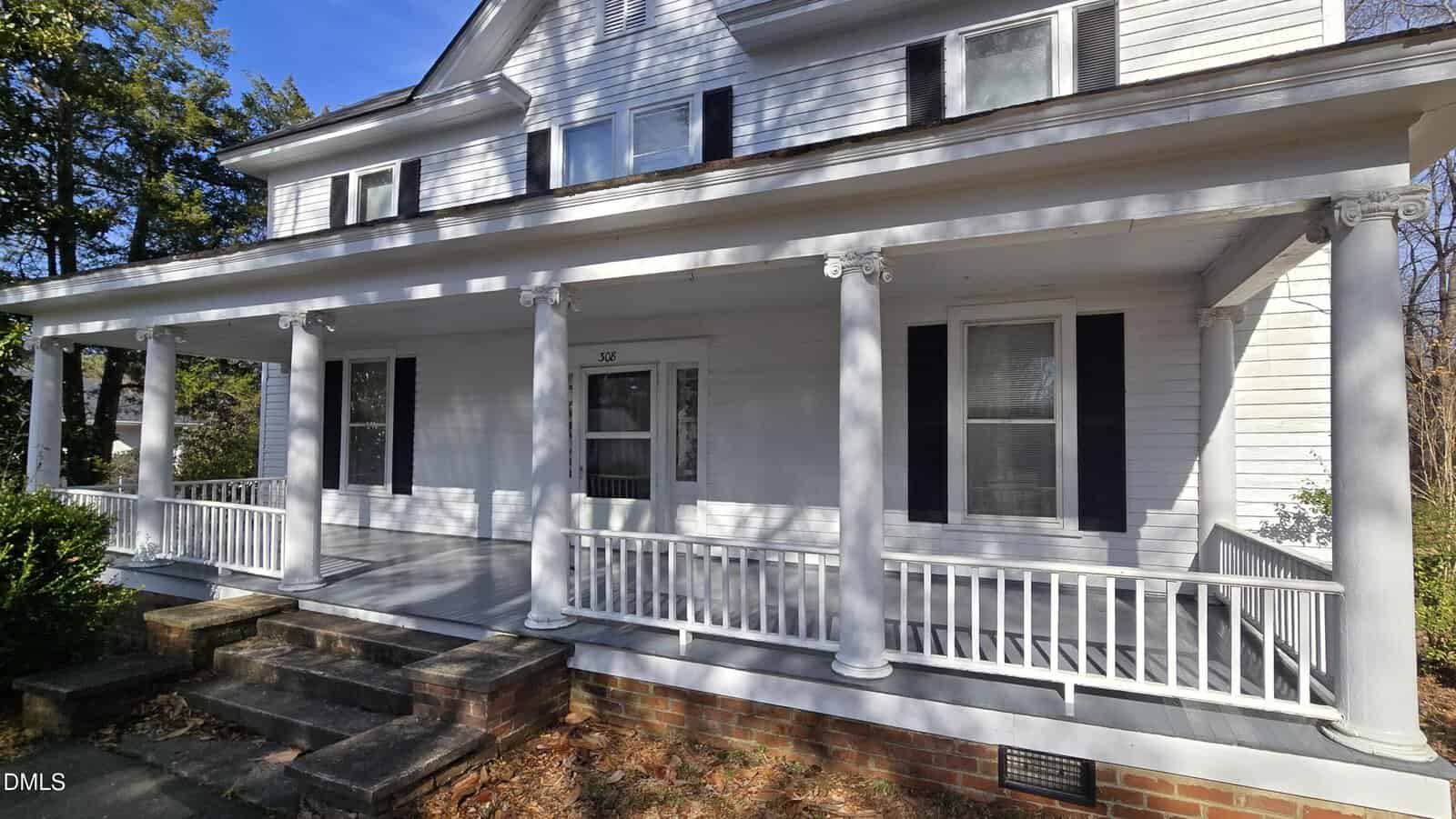 White two-story house with black shutters, a covered front porch supported by white columns, white railing, brick steps, and a screen door. Trees and bushes are visible to the left. The sky is clear and blue.