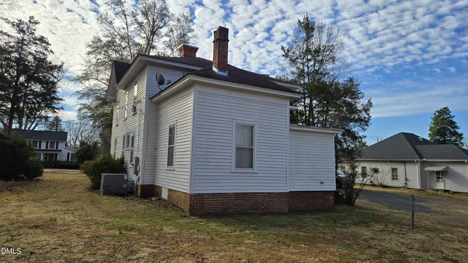 Two-story white house with horizontal siding, brick foundation, and multiple chimneys, seen from the side. The yard is covered with dry grass. Leafless trees and neighboring houses are visible in the background under a cloudy sky.